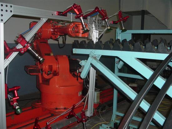 A red industrial robot arm in a work cell with pneumatic clamps and a toothed conveyor.